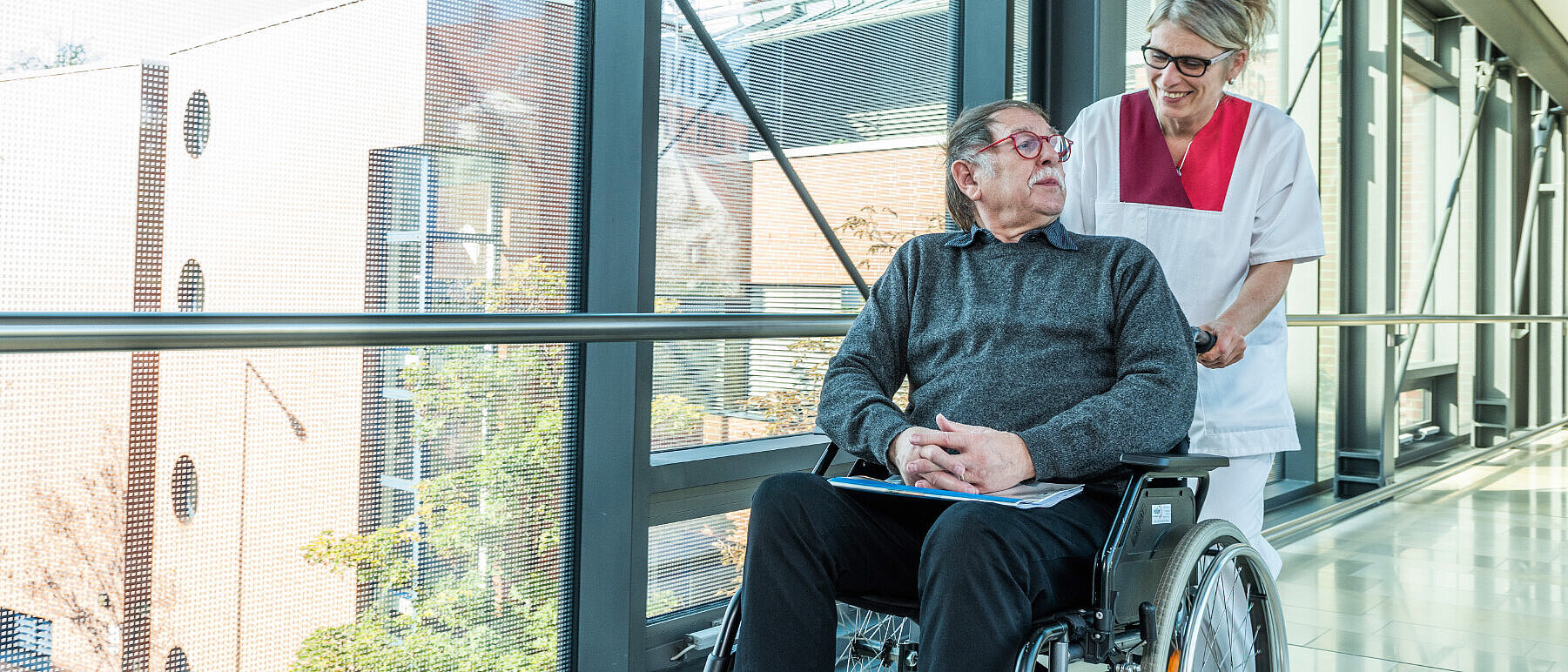 Eine Pflegerin begleitet einen Patienten im Rollstuhl von der Erler-Klinik durch eine Glasbrücke zur Erler-Reha.